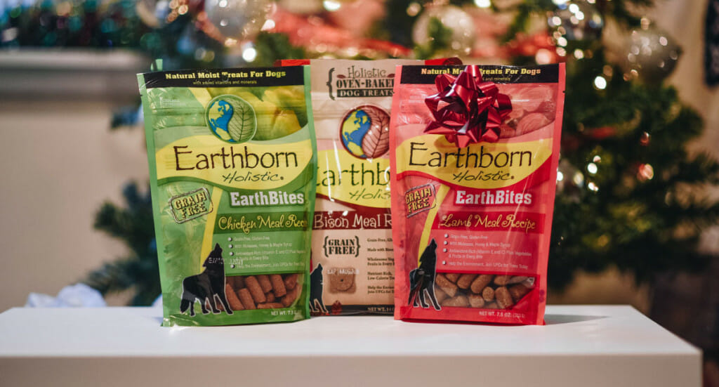 A group of three Earthborn Holistic dog treats sit on a table in front of a Christmas tree, one with a bow on it